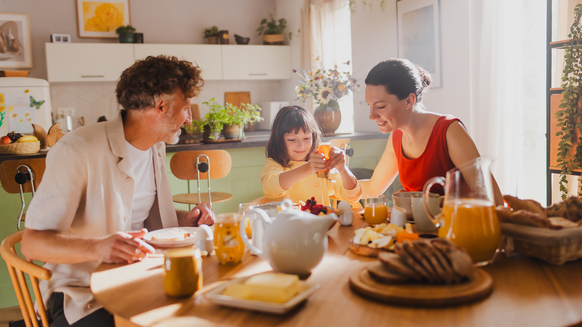 Familie sitzt am Tisch und das Kind macht sich Honig aus dem Spender auf die Pancakes.
